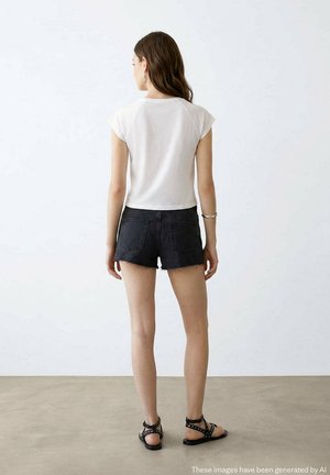 Donna di spalle che indossa una maglietta bianca con maniche corte, pantaloncini di jeans neri, sandali neri con borchie, in piedi davanti a uno sfondo chiaro e uniforme.
