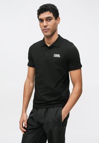 Čierna polokošeľa z bavlnenej zmesi, s golierovým výstrihom a krátkymi rukávmi, s bielym logom potlačeným na ľavej strane hrudníka.