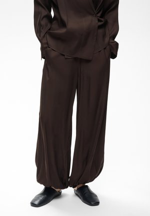 Personne portant un pantalon ample marron foncé avec des poignets élastiques et une chemise assortie, associée à des chaussures noires sans lacets, debout sur un fond blanc.