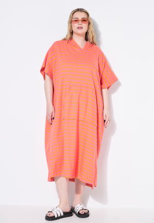 Vestido de rayas, oversized, en rosa brillante y amarillo. Tejido suave, mangas cortas y un bolsillo frontal. Combinado con sandalias tipo slide negras.