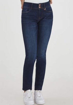 Jeans en denim bleu foncé avec une coupe slim, dotés de deux poches avant, de coutures contrastées et d'une fermeture à deux boutons. Portés avec des baskets blanches.