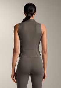 Vrouw met een donkere paardenstaart, gekleed in een mouwloze, olijfgrijs aansluitende top en bijpassende legging, gefotografeerd van achteren tegen een neutrale achtergrond.