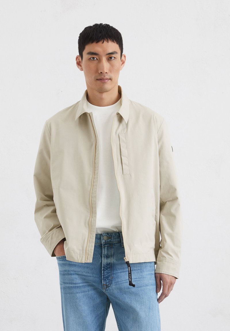 Beige zip-up jacket with a spread collar, flap pocket, and elastic cuffs, paired with a white shirt and light blue jeans.