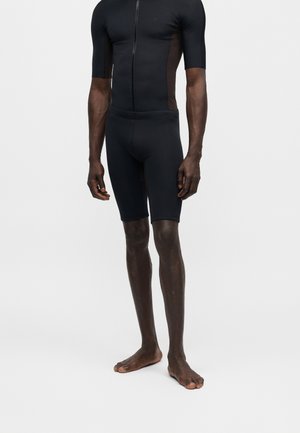 Combinaison de cyclisme noire ajustée avec manches courtes et shorts portée par une personne pieds nus se tenant devant un fond uni.