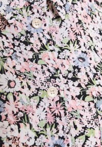 Gros plan sur une chemise à motif floral rose clair, blanc et vert, avec des boutons crème et un col pointu.