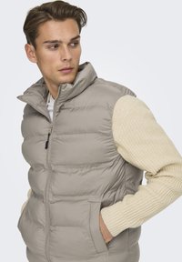 Gilet imbottito in beige chiaro con colletto alto e zip anteriore. Presenta tasche laterali e maniche di un maglione texturizzato color crema.