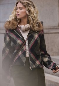 Eine übergroße karierte Jacke in Dunkelgrün, Schwarz und Pink mit Pelzkragen, Reißverschluss und zwei Fronttaschen. Strukturierter Wollstoff.