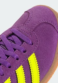 Fioletowy zamszowy sneaker z żółtymi paskami w zygzak, z fioletowymi sznurówkami i brązową gumową podeszwą. Zbliżenie podkreślające teksturę.