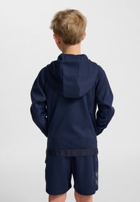 Marinblå huvtröja med dragsko i huvan, långa ärmar och en texturerad nederkant, kombinerad med matchande shorts med gråa chevron-accenter.