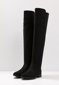 Un par de botas altas de ante negro con tacones bajos, de pie sobre una superficie blanca contra un fondo gris claro.