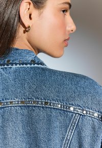 Chaqueta de denim con tachuelas plateadas en los hombros, cuello clásico y un color azul desgastado con un acabado texturizado.