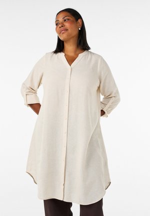 Kvinde med mellem hudtone og mørkt hår iført en lang beige knap-up skjortekjole med op-rullede ærmer mod en ensfarvet hvid baggrund.
