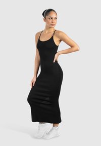 Vestido midi negro ajustado con tirantes finos y escote en forma de u. Llevado con zapatillas blancas, presenta una tela suave y una silueta alargada.
