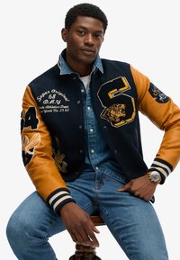 Veste varsity bleu marine et orange avec des patches brodés, y compris un tigre et des chiffres. Design ajusté avec des poignets et un col à rayures côtelées.