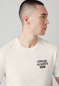 Tricou alb din bumbac cu un imprimeu grafic negru care prezintă textul "HOUSE PLANT'S" și "LEVI'S" în zona stângă a pieptului. Mâneci scurte, guler rotund.