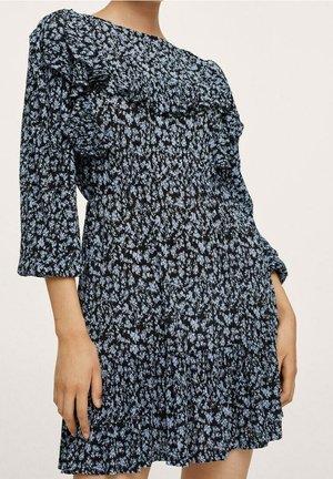 Femme portant une robe noire avec un petit imprimé floral bleu, des manches bouffantes et des détails à volants sur la poitrine et la jupe.