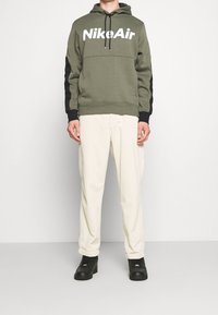 Olivgrön hoodie med vit "NikeAir"-logotyp, svarta detaljer på ärmarna; kombinerad med krämfärgade kordbyxor och svarta sneakers.