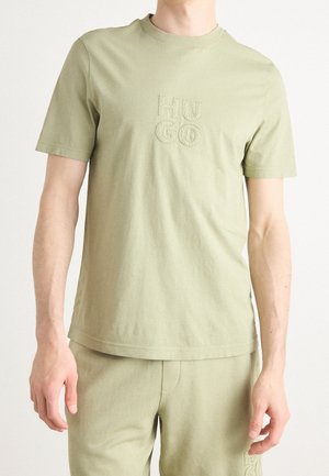 Mężczyzna noszący jasnooliwkowy T-shirt z krótkim rękawem i pasujące spodnie z wytłoczonym napisem "HUGO" na piersi i lewej udzie.