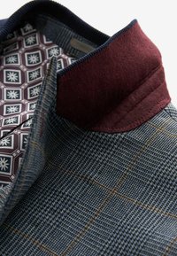 Blazer à carreaux avec un motif bleu marine, col en feutre bordeaux et doublure intérieure à motifs comportant des formes hexagonales et des accents floraux.