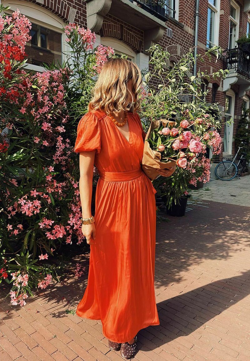 Robe maxi en tissu orange vif, avec des manches bouffantes et une taille cintrée. Tenant un bouquet de roses roses enveloppées dans du papier brun.