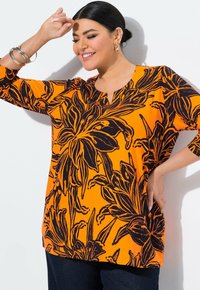 Oranges Oberteil mit schwarzem Blumenmuster, kurzen Ärmeln und einem Schlüssel-Ausschnitt, kombiniert mit dunklen Denim-Hosen. Glatte Stofftextur.