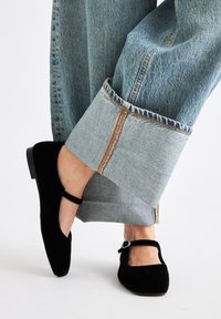 Zapatos Mary Jane de terciopelo negro con un detalle de correa y hebilla, combinados con unos vaqueros de pierna ancha de color azul claro con un dobladillo vuelto y costuras en contraste.