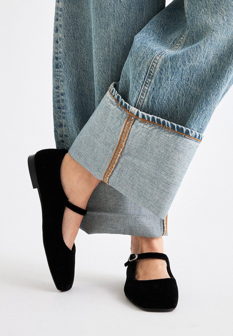 Zapatos Mary Jane de terciopelo negro con un detalle de correa y hebilla, combinados con unos vaqueros de pierna ancha de color azul claro con un dobladillo vuelto y costuras en contraste.