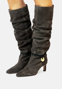 Botas holgadas hasta la rodilla en ante gris oscuro con punta afilada y un tacón cuadrado bajo. Presentan un detalle dorado en el tobillo.