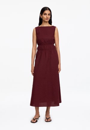 Femme portant une robe midi bordeaux texturée sans manches avec une taille élastique, debout, mains le long du corps, sur fond blanc.