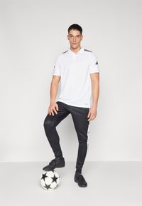 Polo shirt blanc à manches courtes, col à deux boutons et accents noirs, associé à un pantalon de sport noir avec des bandes blanches sur les côtés. Ballon de football noir.