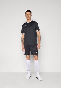 Schwarzes Sporttrikot und -shorts mit weißen Akzenten, mit einem Logo auf der Brust. Kombiniert mit weißen knielangen Socken und Fußballschuhen.