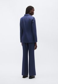 Individualizuotas tamsiai mėlynas kostiumas su aptemptu švarku ir platėjusiomis kelnėmis, pagamintas iš tekstūruotos medžiagos. Nugaros vaizde matyti subtilios detalės ir švarūs kontūrai.