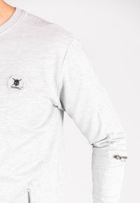 Sudadera gris claro de manga larga, con un parche redondo que presenta un diseño de calavera y un detalle con cremallera en la manga izquierda.