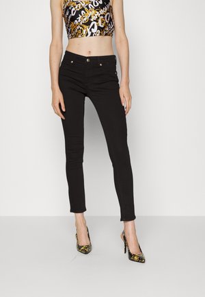 Femme portant un pantalon slim noir, un crop top à motifs et des talons hauts noirs avec un motif floral doré, debout sur fond blanc.