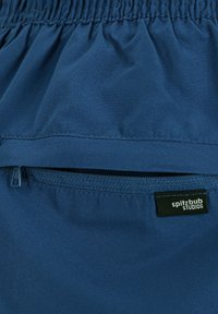 Marineblaues Gewebe mit einem gerafften Bund, einer Reißverschlusstasche und einem Etikett mit der Aufschrift "spitzbub STUDIOS." Glatte Textur, matte Oberfläche.