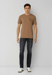 Braunes T-Shirt mit kurzen Ärmeln und einer Taschen-Grafik, kombiniert mit dunklen Jeans und weißen Turnschuhen. Glatte Textur und lässige Passform.