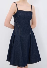 Jeanskleid mit Spaghettiträgern, quadratischem Ausschnitt und ausgestelltem Rock. Dunkelblauer Stoff mit goldenen Nähten und zwei seitlichen Taschen.