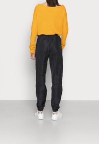 Přes oversized žlutý pletený svetr, černé saténové tepláky s elastickými manžetami a bílé tenisky s černými akcenty. Měkká textura, uvolněný střih.