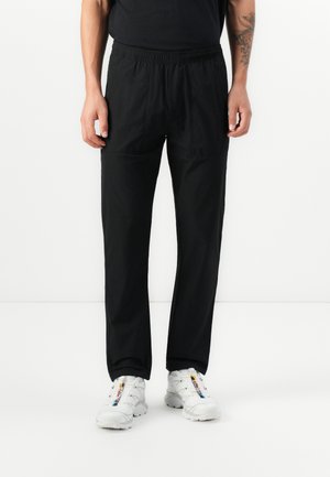 Black athletic pants with an elastic waist, two side pockets, straight legs, and a lightweight material, worn with white sneakers.