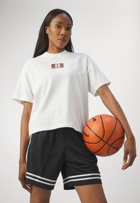 White cropped t-shirt with a maroon logo, paired with black shorts featuring white stripes. The figure holds an orange basketball.