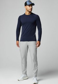 Marineblaue Langarmshirt mit strukturierten Seiten, hellgraue Stretchhose und weiße Sportschuhe. Blaue und schwarze Mütze mit Logo-Akzenten.