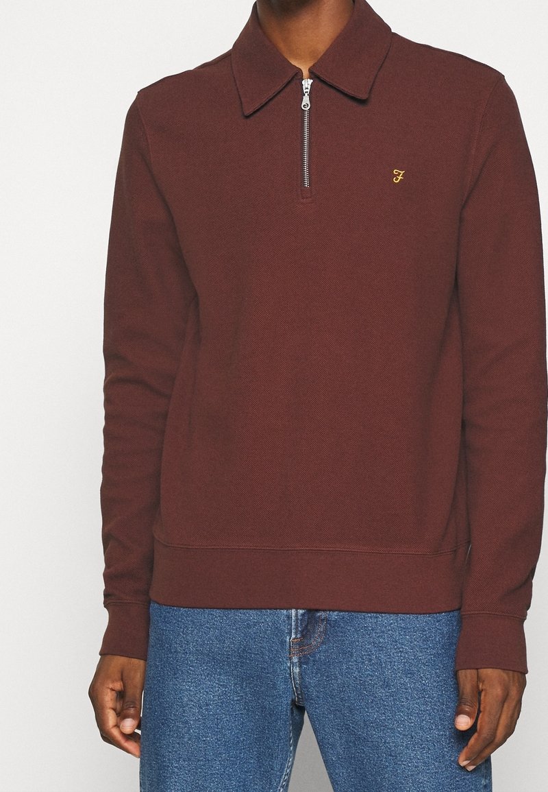Homme portant un pull rouge foncé à manches longues avec une fermeture éclair devant et un petit F jaune brodé sur la poitrine, associé à un jean bleu.