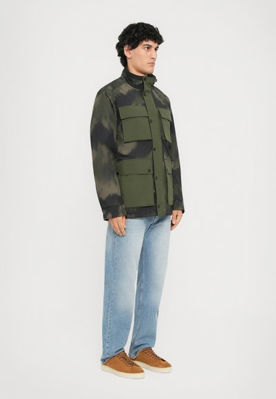 Kamuflagejakke i mørkegrøn og sort, med høj krave, store forlommer og knaplukning, parret med lyseblå jeans og brune sko.