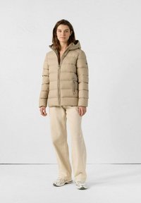 Frau steht gegen eine weiße Wand, trägt eine beige Pufferjacke mit Kapuze, cremefarbene Hose und silberne Sneaker, schaut in die Kamera, Hände entspannt.