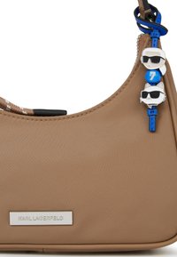 Pruun nailonnahast käekott kõveriku disainiga, millel on tekstuursed detailid ja värvikad ripatsid, sealhulgas multifilmitegelased ja sinine helm.