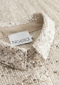 Kreminės spalvos blizgučiais puoštas marškinėlių apykaklė su dviem baltais mygtukais ir matoma "Noella" prekės ženklo etikete apykaklės viduje.