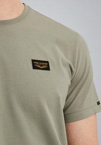 Olive green cotton t-shirt with short sleeves, featuring a black rectangular patch with gold lettering and a small logo detail.
