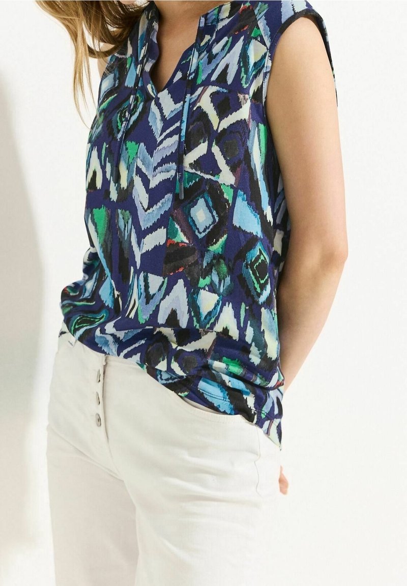 Ärmlös topp i djupblått med abstrakta geometriska mönster i grönt, svart och vitt. Tyget verkar lätta; har en v-ringning.