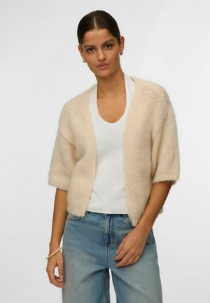 Nő krémszínű kötött kardigánban, fehér póló és világoskék farmer fölött, sima háttér előtt áll.