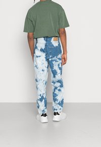 Sinivalkoiset solmitut farkut, joissa on valkoisia pilvikuoseja ja vaaleanruskea pala takataskussa, yhdistettynä vihreään ylisuurikokoiseen T-paitaan ja valkoisiin lenkkareihin.